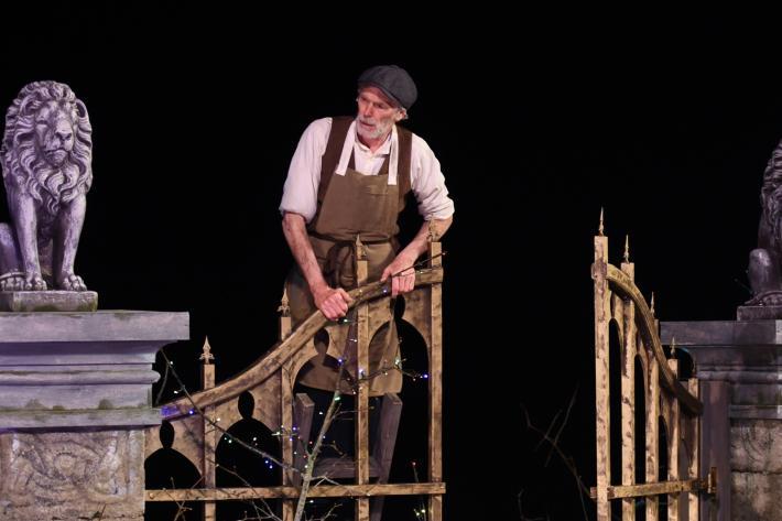 An actor on a ladder peers over an iron gate