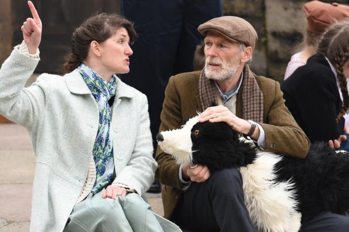 An old man sits on the stage with a life size collie puppet, talking to a woman who is gesticulating